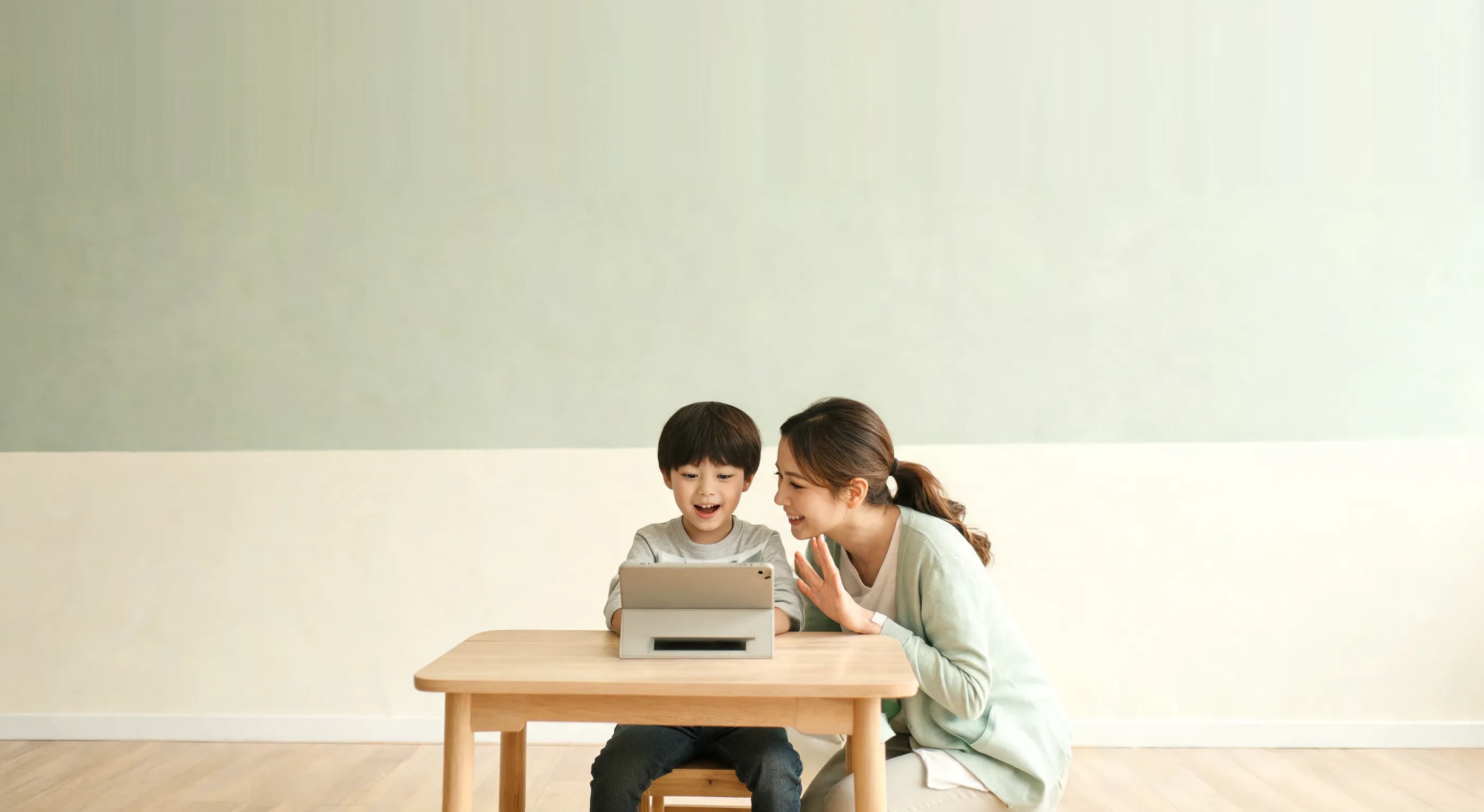 A speech therapist leaning in beside a young child who is speaking aloud to a tablet during a guided practice activity